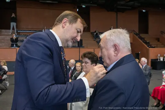 Mężczyzna przypina odznaczenie do marynarki innego mężczyzny w sali pełnej ludzi.