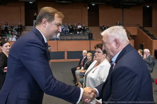 Mężczyźni uściskiem dłoni podczas ceremonii wręczenia odznaczeń. W tle siedzą goście.