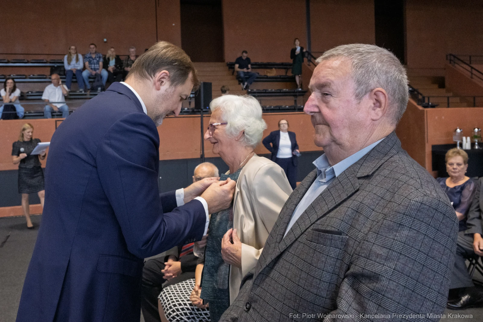 Wręczenie odznaczenia starszej kobiecie przez mężczyznę w garniturze, obok stoi inny mężczyzna.  Autor: Piotr Wojnarowski