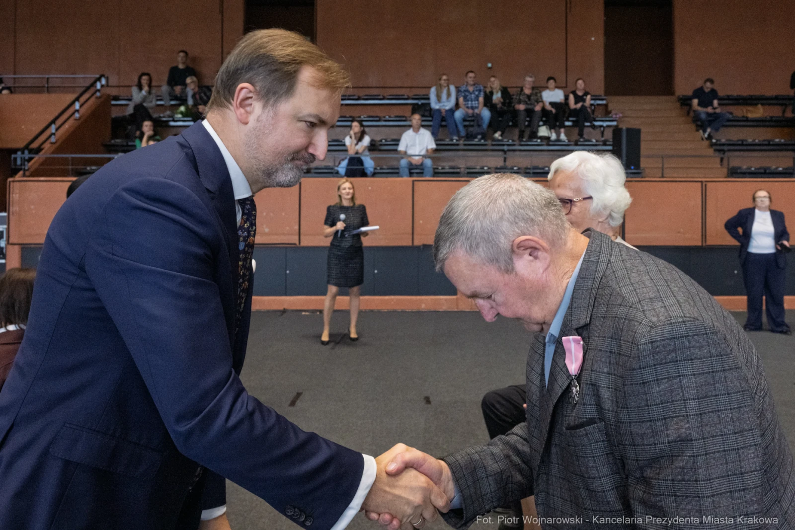 Mężczyzna w garniturze podaje rękę starszemu mężczyźnie w eleganckim ubraniu. W tle widoczna publiczność.  Autor: Piotr Wojnarowski