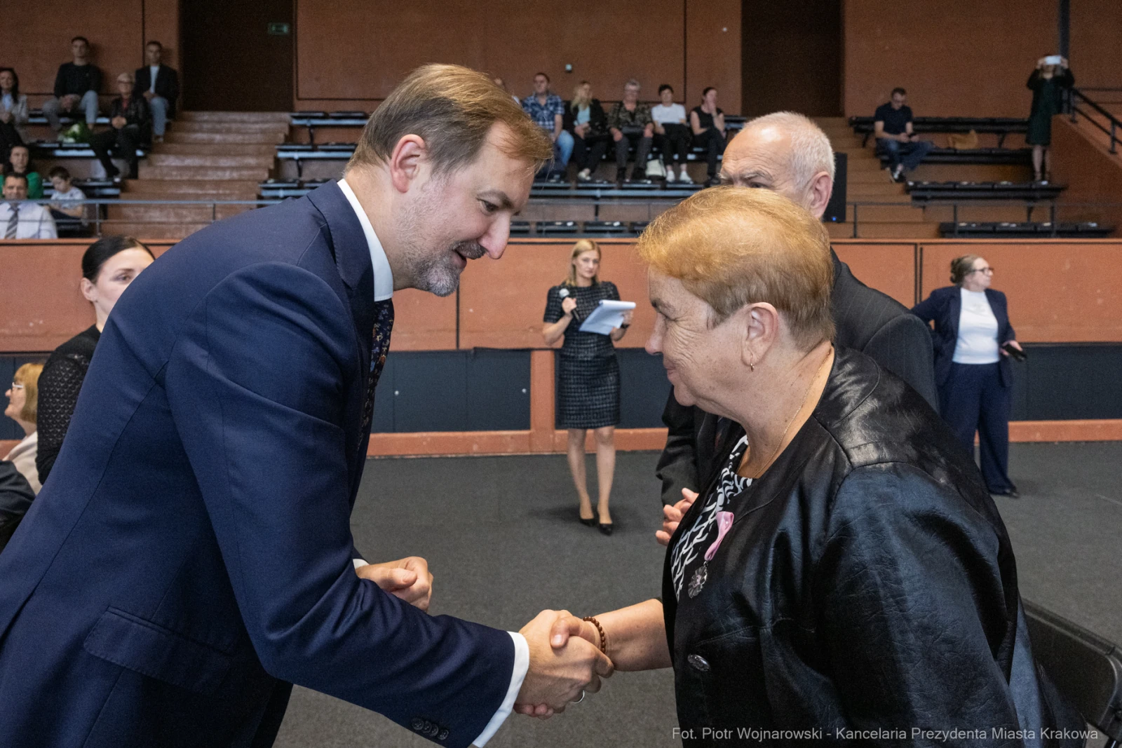 Mężczyzna uścisnął dłoń kobiecie w formalnym otoczeniu, w tle widoczni inni uczestnicy wydarzenia.  Autor: Piotr Wojnarowski