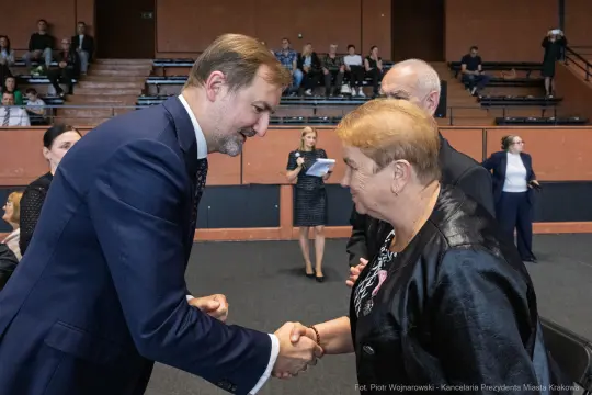 Mężczyzna uścisnął dłoń kobiecie w formalnym otoczeniu, w tle widoczni inni uczestnicy wydarzenia.
