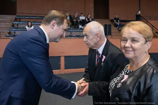 Uścisk dłoni między mężczyzną a starszym mężczyzną, obok kobieta w jasnym ubraniu.