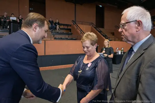 Wręczenie odznaczenia przez mężczyznę kobiecie, obok stoi starszy mężczyzna.
