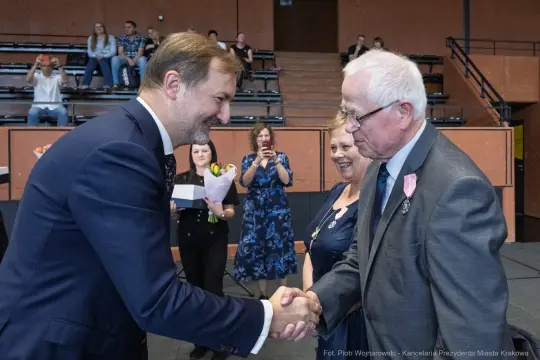 Uścisk dłoni dwóch mężczyzn podczas uroczystości, w tle goście z kwiatami.