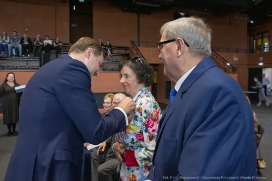 Mężczyzna przypina odznaczenie kobiecie w kolorowej sukience, obok stoi mężczyzna w okularach.