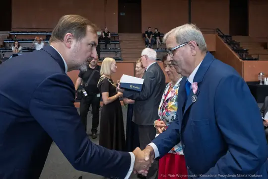 Uścisk dłoni dwóch mężczyzn podczas ceremonii wręczenia nagród. W tle widoczni inni uczestnicy.