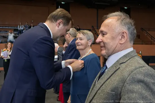 Wręczenie odznaczeń przez mężczyznę starszej kobiecie w towarzystwie innych osób.