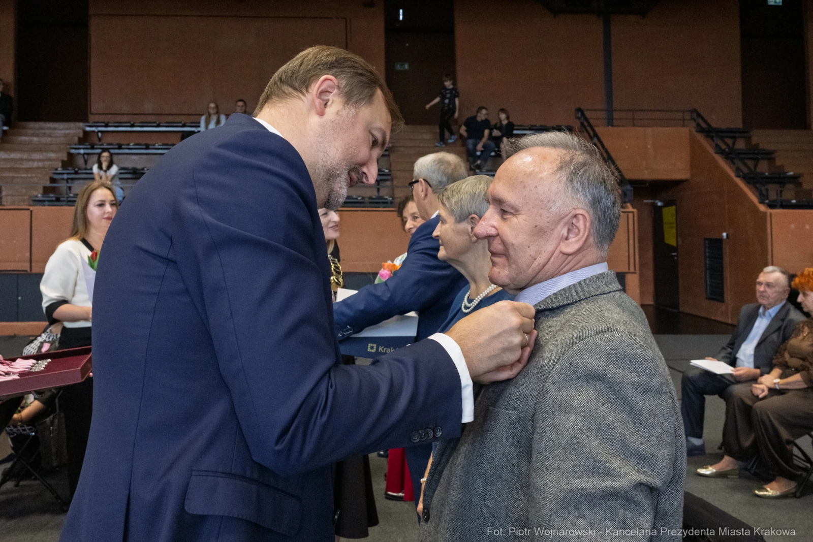 Wręczenie odznaczenia przez mężczyznę w garniturze innemu mężczyźnie w szarym ubraniu.  Autor: Piotr Wojnarowski