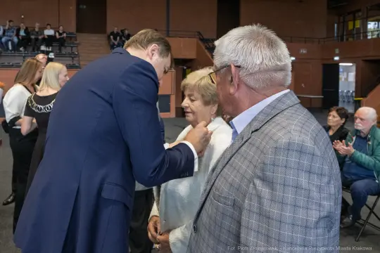Wręczenie odznaczenia przez mężczyznę starszej kobiecie w obecności innych gości.