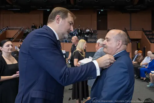 Wręczenie odznaczenia mężczyźnie przez innego mężczyznę w formalnej atmosferze.
