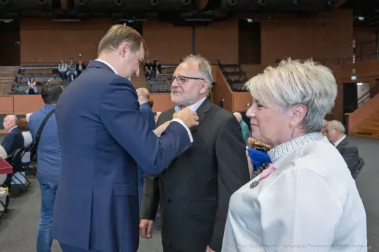 Mężczyzna przypina odznaczenie innemu mężczyźnie w obecności kobiety.