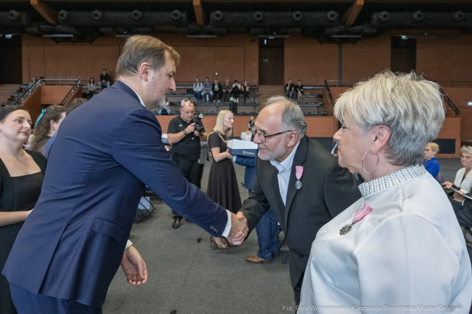 Uścisk dłoni między mężczyzną a starszym mężczyzną w formalnej scenerii. W tle widoczna publiczność.  Autor: Piotr Wojnarowski