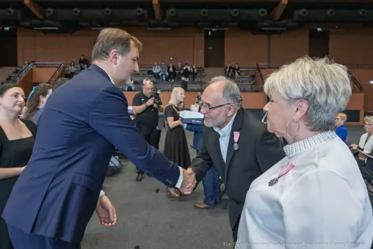 Uścisk dłoni między mężczyzną a starszym mężczyzną w formalnej scenerii. W tle widoczna publiczność.