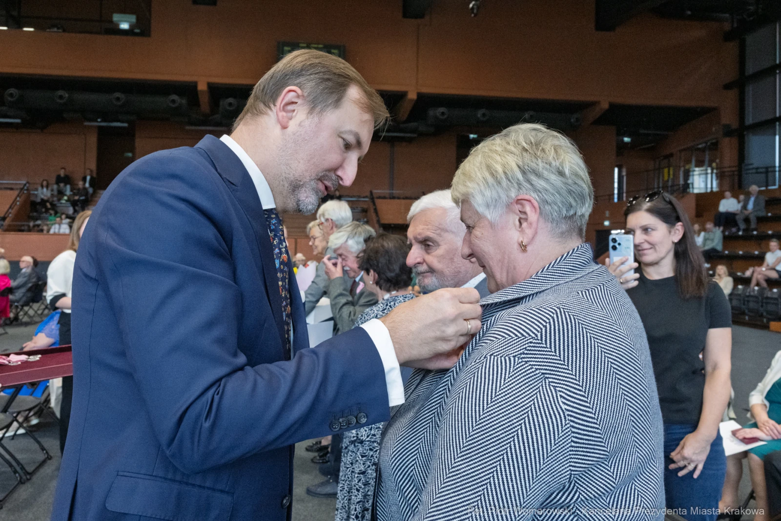 Mężczyzna przypina odznaczenie do ubrania kobiety w towarzystwie innych osób.  Autor: Piotr Wojnarowski