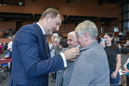 Mężczyzna przypina odznaczenie do ubrania kobiety w towarzystwie innych osób.