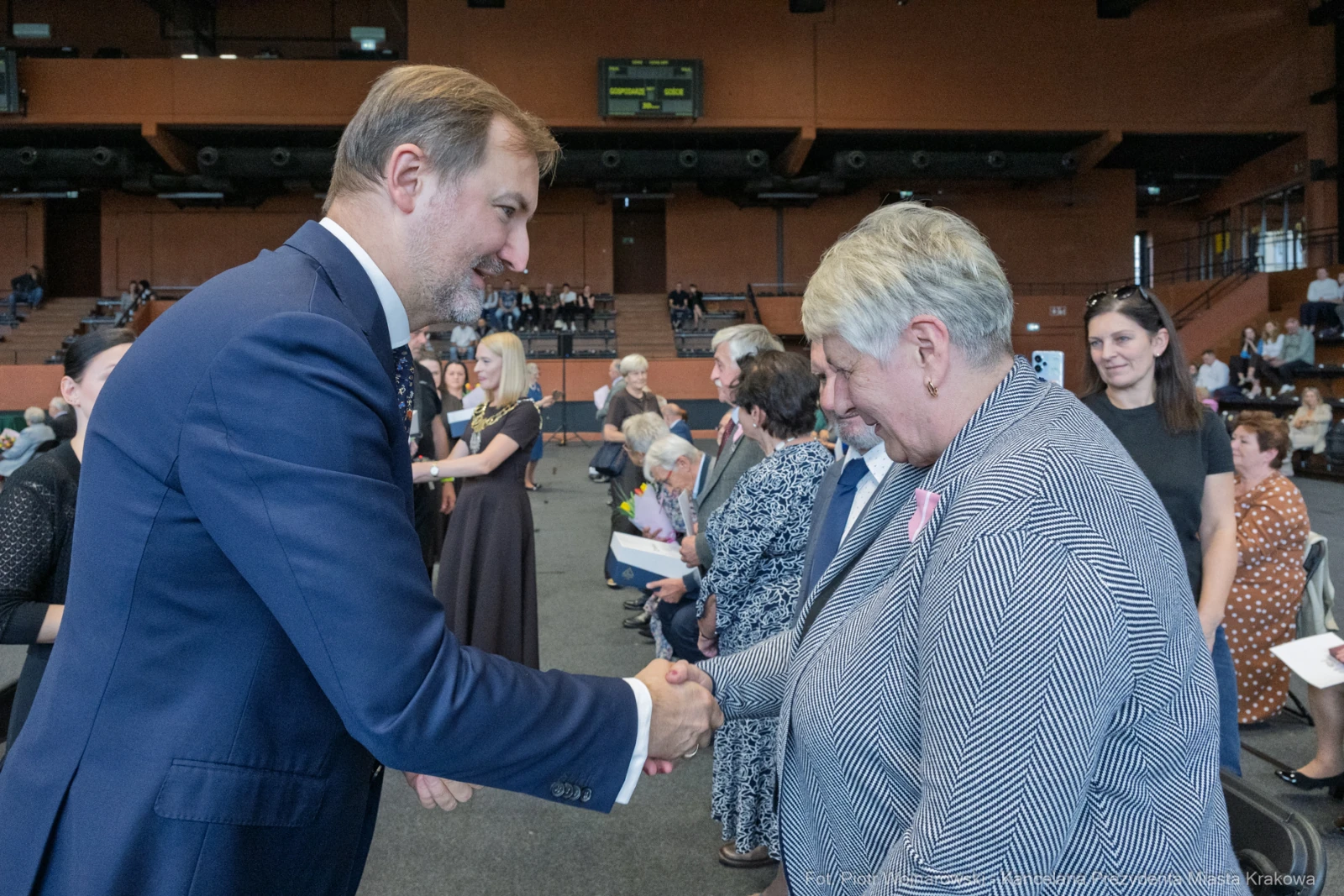 Uścisk dłoni między mężczyzną a kobietą w formalnej atmosferze. W tle zgromadzenie ludzi.  Autor: Piotr Wojnarowski
