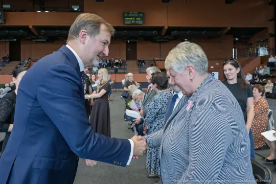 Uścisk dłoni między mężczyzną a kobietą w formalnej atmosferze. W tle zgromadzenie ludzi.