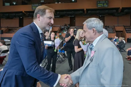 Mężczyzna w garniturze uścisnął dłoń starszemu mężczyźnie, w tle inni uczestnicy wydarzenia.