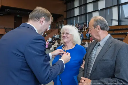 Wręczenie odznaczenia kobiecie przez mężczyznę w eleganckim otoczeniu. Dwoje osób obserwuje moment.
