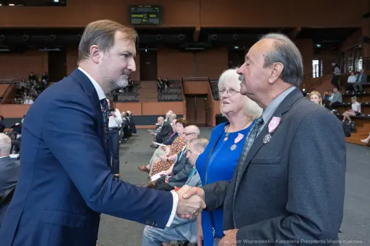 Mężczyzna w garniturze uścisnął dłoń starszemu mężczyźnie, obok stoi kobieta w okularach.