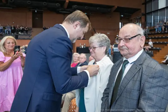 Uroczystość wręczenia odznaczeń, osoby w eleganckich strojach, aplauz zgromadzonych.
