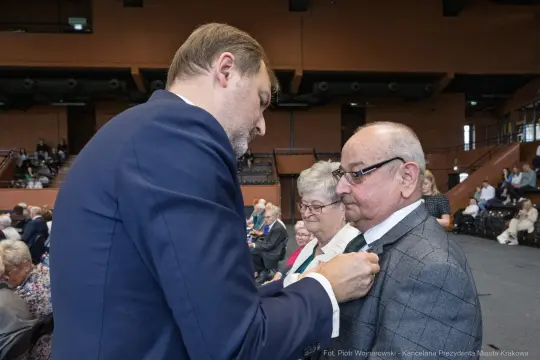 Mężczyzna przypina odznaczenie starszemu mężczyźnie w towarzystwie kobiety.