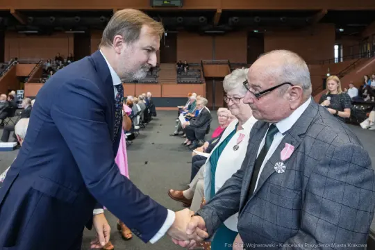 Mężczyzna w garniturze podaje rękę starszemu mężczyźnie z odznaczeniem, w tle siedzą inni goście.