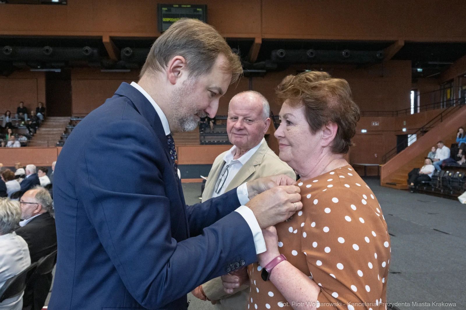 Wręczenie odznaczenia przez mężczyznę w garniturze kobiecie w brązowej sukience w kropki. W tle mężczyzna.  Autor: Piotr Wojnarowski