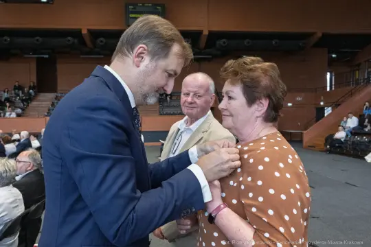 Wręczenie odznaczenia przez mężczyznę w garniturze kobiecie w brązowej sukience w kropki. W tle mężczyzna.