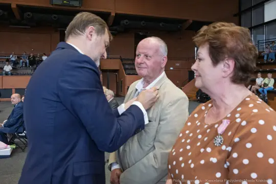 Mężczyzna odbiera odznaczenie od drugiego mężczyzny, obok stoi kobieta.
