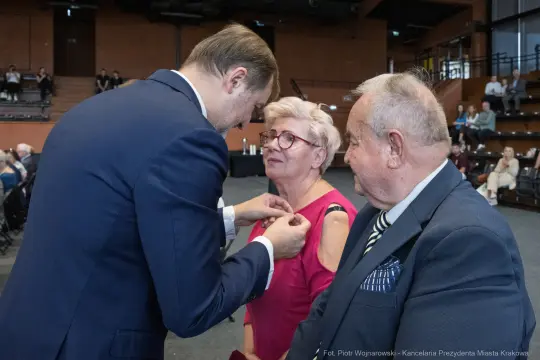 Uroczystość wręczenia odznaczenia kobiecie przez mężczyznę w garniturze. Obok starszy mężczyzna.