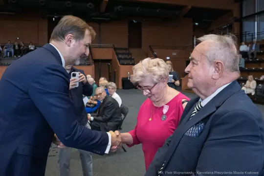 Spotkanie dwóch starszych osób z mężczyzną w garniturze, uściski dłoni i odznaczenia.
