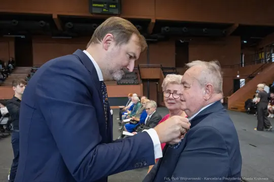 Wręczenie odznaczenia mężczyźnie przez elegancko ubranego mężczyznę w towarzystwie starszej pary.