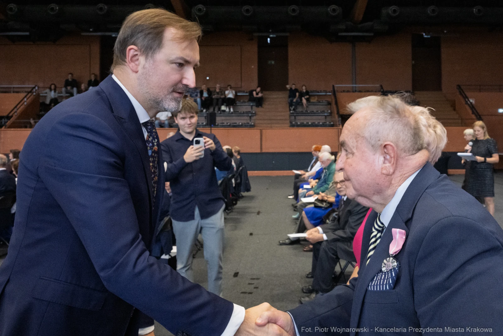 Mężczyzna w garniturze uścisnął dłoń starszemu mężczyźnie w eleganckim stroju. W tle widoczna publiczność.  Autor: Piotr Wojnarowski