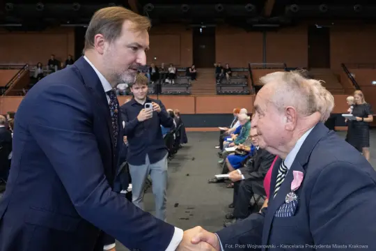 Mężczyzna w garniturze uścisnął dłoń starszemu mężczyźnie w eleganckim stroju. W tle widoczna publiczność.