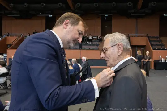Mężczyzna przypina odznaczenie do marynarki innego mężczyzny podczas uroczystości.