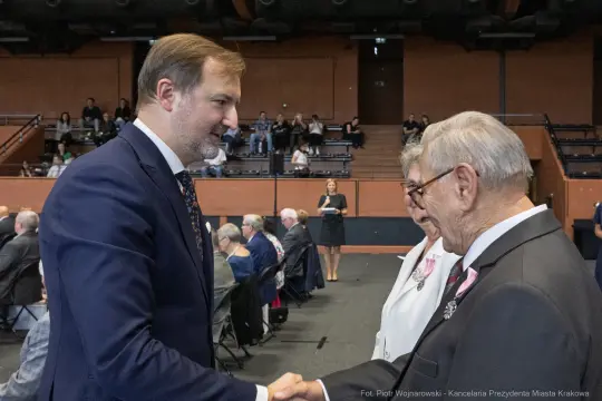 Uścisk dłoni między mężczyzną a starszym panem, w tle widoczna grupa ludzi.