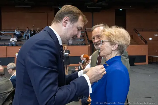 Wręczenie odznaczenia przez mężczyznę kobiecie w niebieskiej bluzce, w tle widownia.