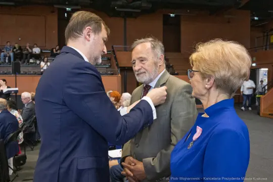 Mężczyzna przypina odznaczenie do marynarki innego mężczyzny, obok stoi kobieta.