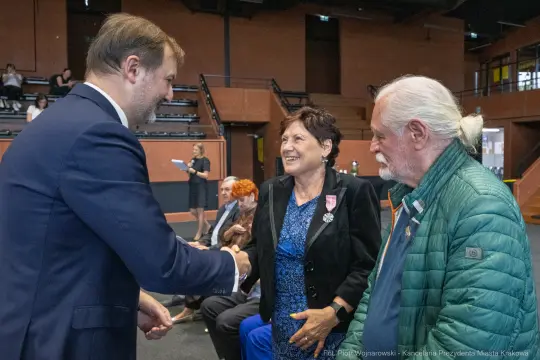 Uścisk dłoni między mężczyzną a kobietą, obok starszy mężczyzna z białymi włosami.