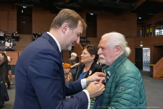 Mężczyzna przypina odznaczenie starszemu mężczyźnie w towarzystwie innych osób.