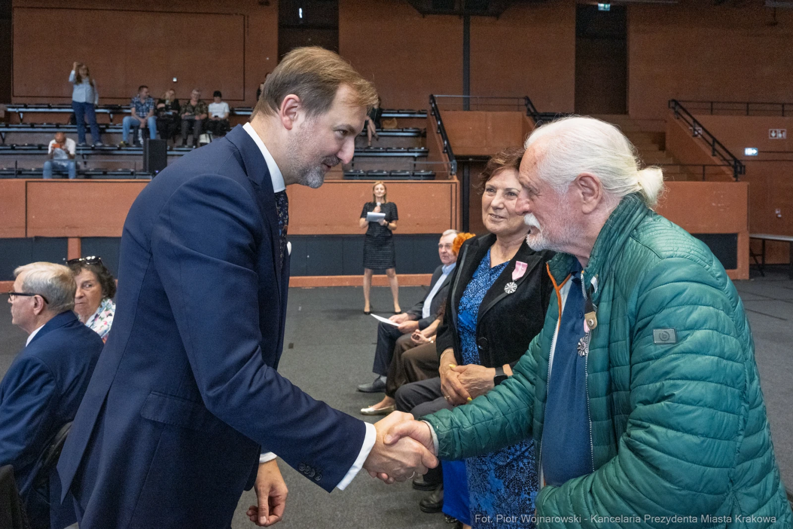 Uroczystość wręczenia odznaczeń, mężczyzna w garniturze podaje rękę starszemu mężczyźnie.  Autor: Piotr Wojnarowski