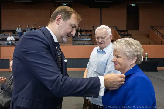 Wręczenie odznaczenia przez mężczyznę kobiecie, w tle starszy mężczyzna.
