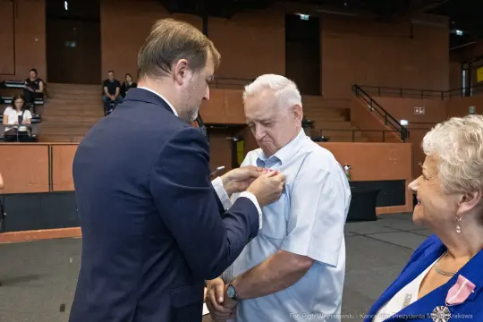 Mężczyzna odbiera odznaczenie, obok uśmiechająca się kobieta.