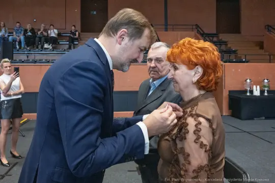 Wręczenie odznaczenia przez mężczyznę z brązowym garniturem kobiecie z rudymi włosami. W tle widownia.