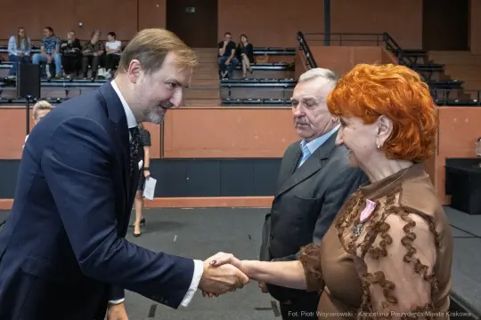 Mężczyzna w garniturze podaje rękę kobiecie z rudymi włosami, w tle inni uczestnicy wydarzenia.