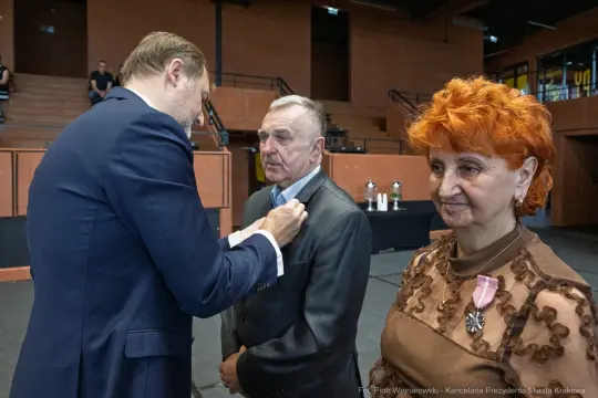 Ceremonia wręczenia odznaczeń, mężczyzna w garniturze, kobieta z pomarańczowymi włosami.
