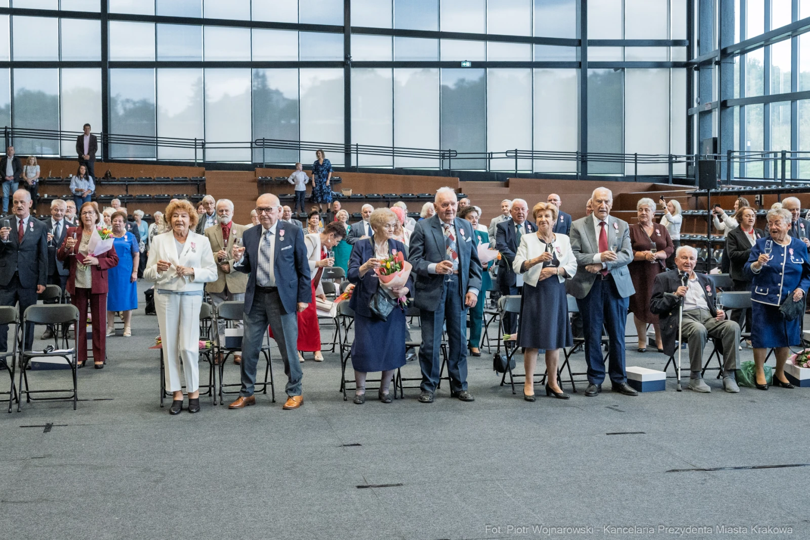 Grupa ludzi stojących w sali, trzymających kwiaty, podczas uroczystości.  Autor: Piotr Wojnarowski