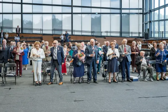 Grupa ludzi stojących w sali, trzymających kwiaty, podczas uroczystości.